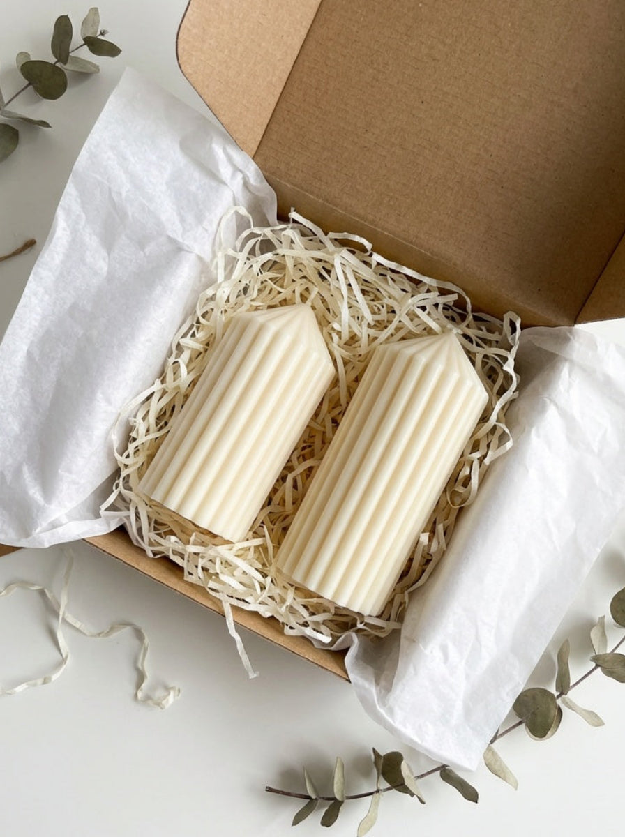 Two white pillar candles in a brown box with white tissue paper on a light surface.

